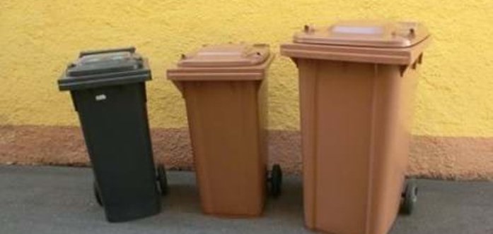 Drei nebeneinander stehende Biotonnen in brauner und grauer Farbe vor einer gelben Wand. Die Tonnen unterscheiden sich in der Größe, vermutlich 120, 240 und 360 Liter.