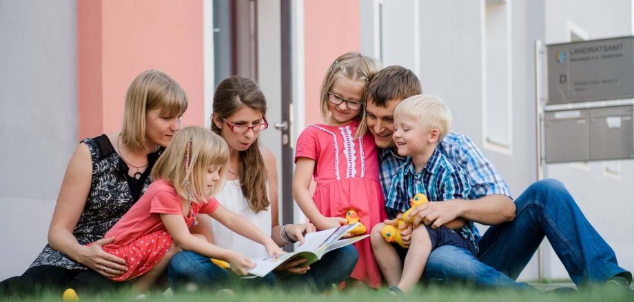 Eine Familie befindet sich eine Wiese und schaut zusammen ein Buch an.