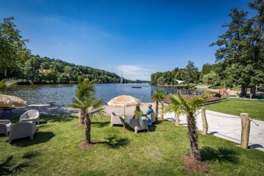 Blick auf ein idyllisches Naturfreibad mit Liegewiese, Palmen, Sonnenschirmen und Sitzgelegenheiten am Ufer eines Sees. Menschen entspannen im Schatten, der See ist von Wald umgeben, im Hintergrund ist ein Steg und ein Wasserfontäne sichtbar. Sommerliche Atmosphäre bei blauem Himmel.