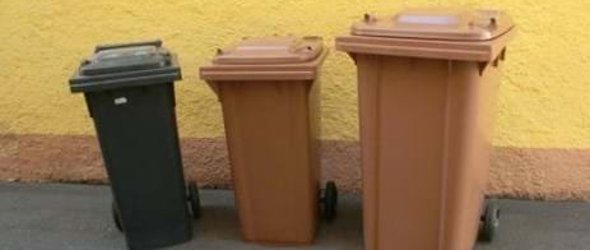 Drei nebeneinander stehende Biotonnen in brauner und grauer Farbe vor einer gelben Wand. Die Tonnen unterscheiden sich in der Größe, vermutlich 120, 240 und 360 Liter.