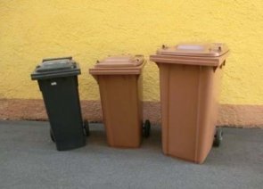 Drei nebeneinander stehende Biotonnen in brauner und grauer Farbe vor einer gelben Wand. Die Tonnen unterscheiden sich in der Größe, vermutlich 120, 240 und 360 Liter.
