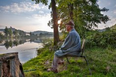 Ein Mann sitzt mit seiner Angel an einem ruhigen Weiher mit idyllischen Hintergrund.