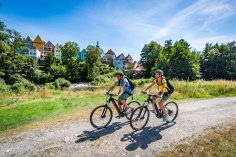 Zwei Radfahrer fahren bei Sonnenschein auf einem Schotterweg durch eine grüne Flusslandschaft. Im Hintergrund bunte Häuser einer historischen Altstadt mit Turm und Bäumen.