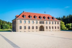 KZ-Gedenkstätte Flossenbürg
© Oberpfälzer Wald, Fotograf Thomas Kujat Gebäude der KZ-Gedenkstätte Flossenbürg aus hellem Sandstein mit rotem Dach, vielen Fenstern und einem zentralen Torbogen. Das Gebäude steht auf einem weitläufigen, hellen Vorplatz mit Blick auf eine grüne Waldlandschaft im Hintergrund.