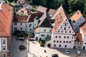 Luftaufnahme einer kleinen Stadt mit historischen und modernen Gebäuden. Im Zentrum steht ein großes Gebäude mit rotem Satteldach und grauer Fassade, daneben ein moderner Gebäudekomplex mit flachem Dach. Rechts schließen sich alte Häuser mit bunten Fassaden und spitzen Giebeldächern an.