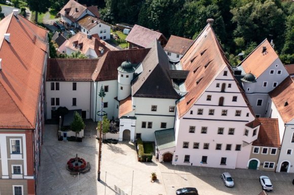 Luftaufnahme einer kleinen Stadt mit historischen und modernen Gebäuden. Im Zentrum steht ein großes Gebäude mit rotem Satteldach und grauer Fassade, daneben ein moderner Gebäudekomplex mit flachem Dach. Rechts schließen sich alte Häuser mit bunten Fassaden und spitzen Giebeldächern an.