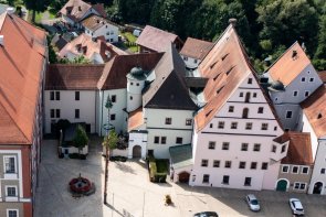 Dienstgebäude Landratsamt Neustadt an der Waldnaab
© Landkreis Neustadt an der Waldnaab Luftaufnahme einer kleinen Stadt mit historischen und modernen Gebäuden. Im Zentrum steht ein großes Gebäude mit rotem Satteldach und grauer Fassade, daneben ein moderner Gebäudekomplex mit flachem Dach. Rechts schließen sich alte Häuser mit bunten Fassaden und spitzen Giebeldächern an.